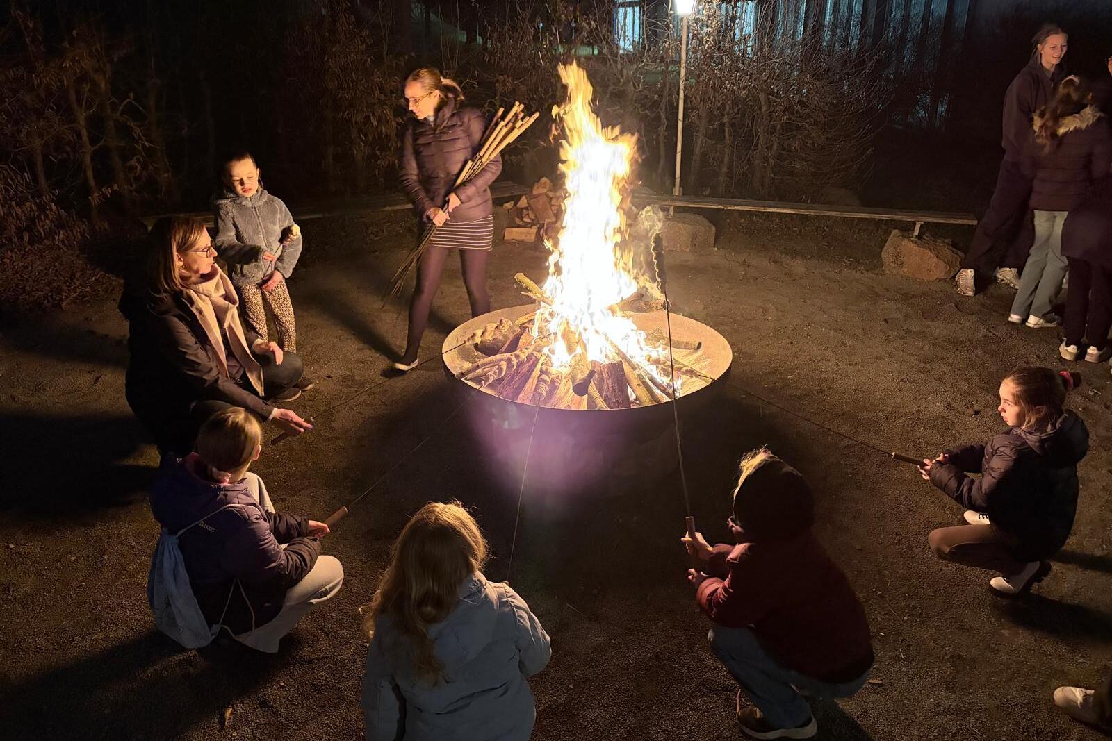 Gruppensitzung um ein Lagerfeuer, Kinder und Erwachsene mit Stockbrot, beleuchtet von Flammen in nächtlicher Umgebung.
