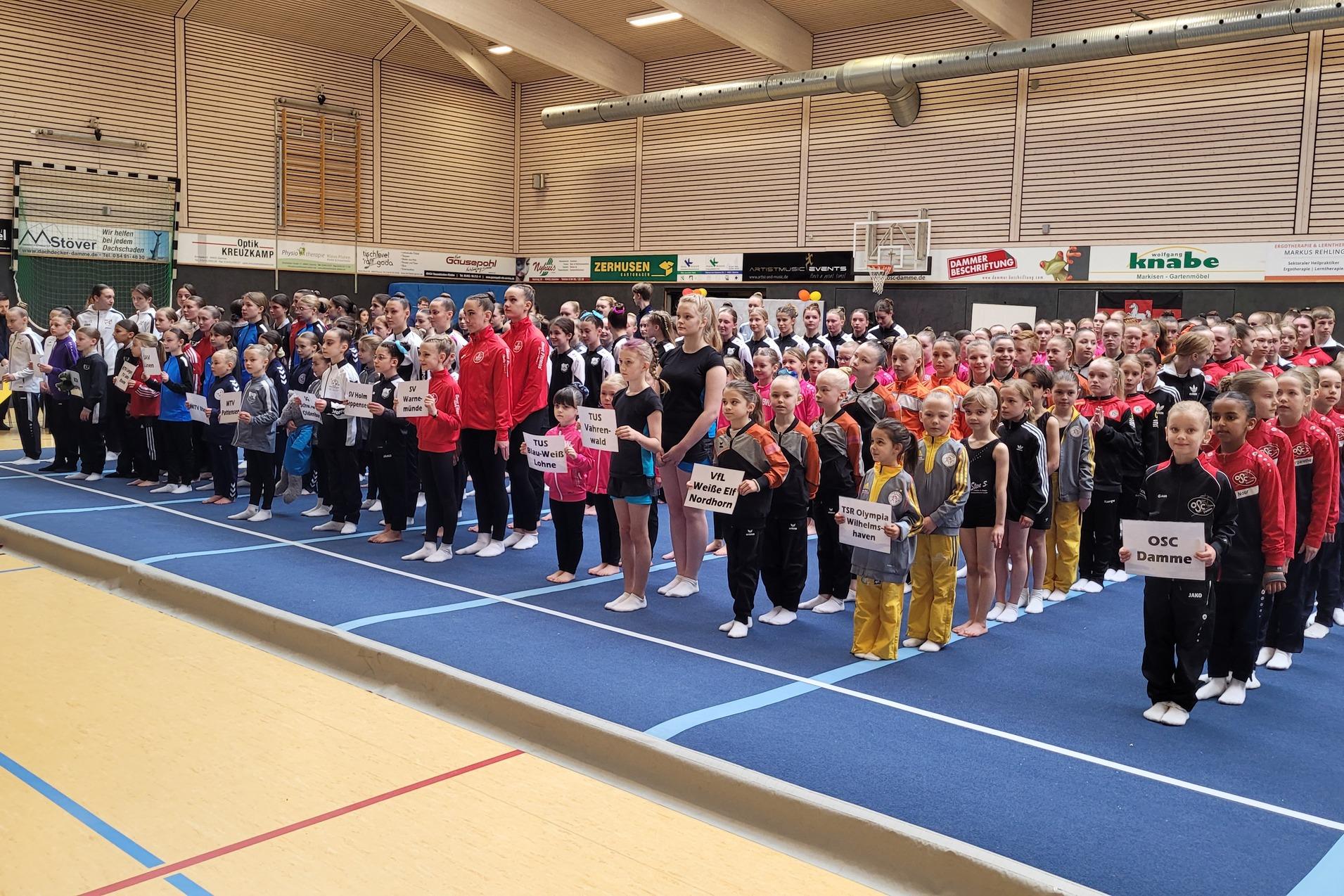 Gruppenfoto von Sportlern in Wettkampfkleidung, versammelt auf einer blauen Turnmatte, mit Namensschildern in den Händen.
